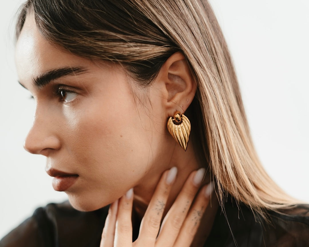 Donna bionda con top nero che tocca delicatamente orecchini dorati Medema minimalisti, rappresentazione della filosofia dell'eleganza come forma di espressione personale